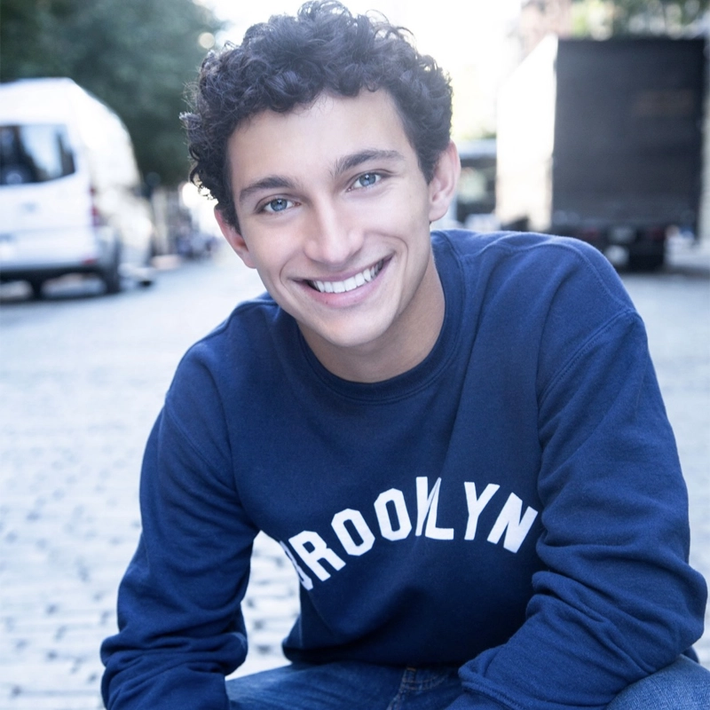 Jonathan Fenton, a young man with curly dark hair and blue eyes, smiles warmly at the camera. He wears a navy blue 