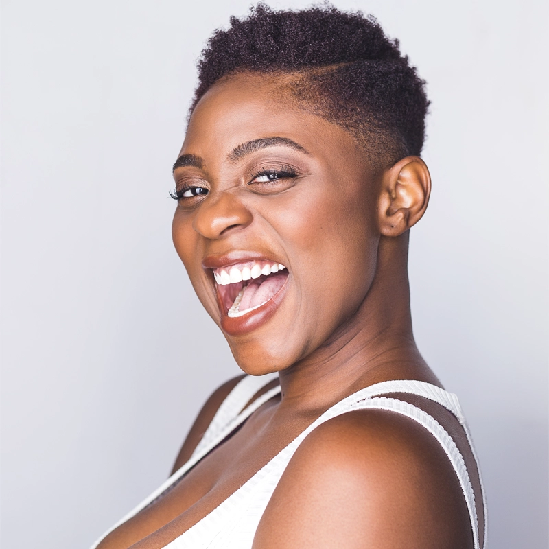 A woman with short natural hair smiles widely, showing her teeth. She is wearing a white sleeveless top and faces the camera at a three-quarter angle against a plain light background. Her expression is joyful and energetic.