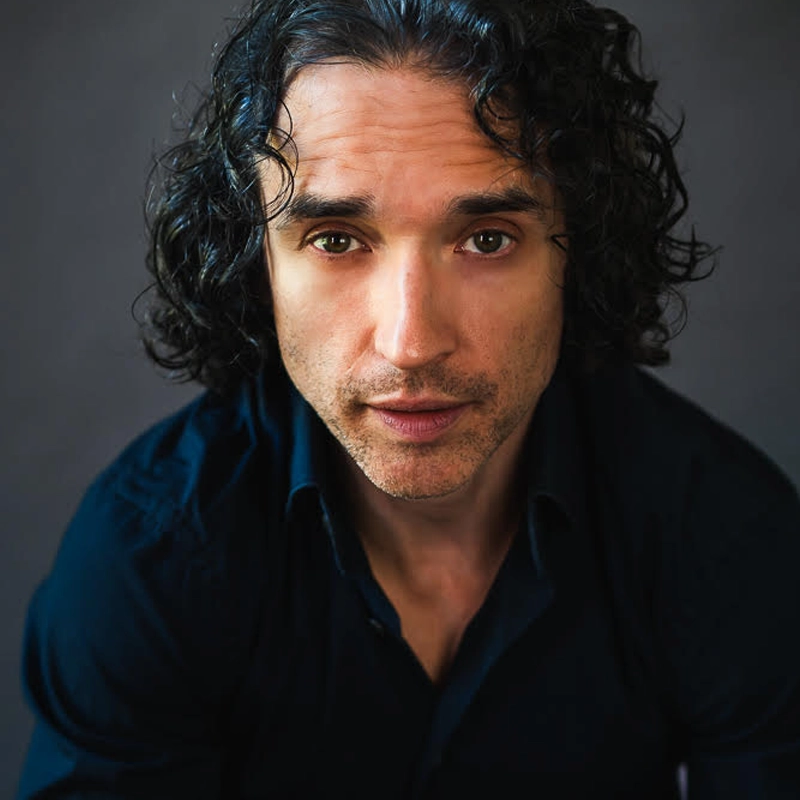A man with medium-length, curly dark hair and light skin gazes directly at the camera. He wears a dark collared shirt and sits against a dark, blurred background. His expression is serious and contemplative.