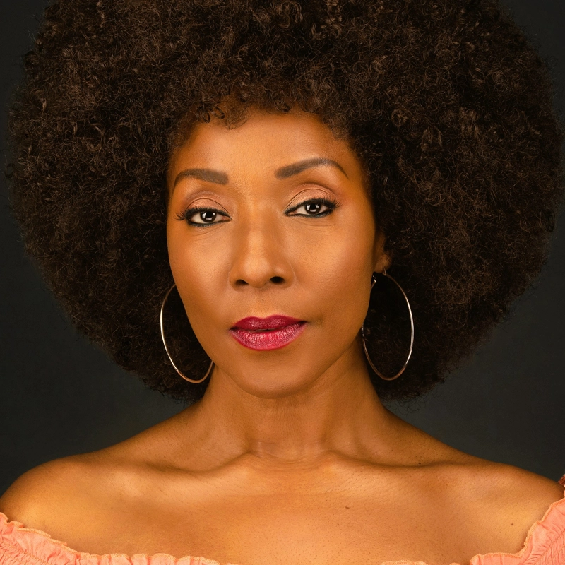 A woman with dark skin and an afro hairstyle, resembling Harriet D. Foy, looks confidently at the camera. She wears large hoop earrings, berry-colored lipstick, and an off-the-shoulder peach top against a striking black background.