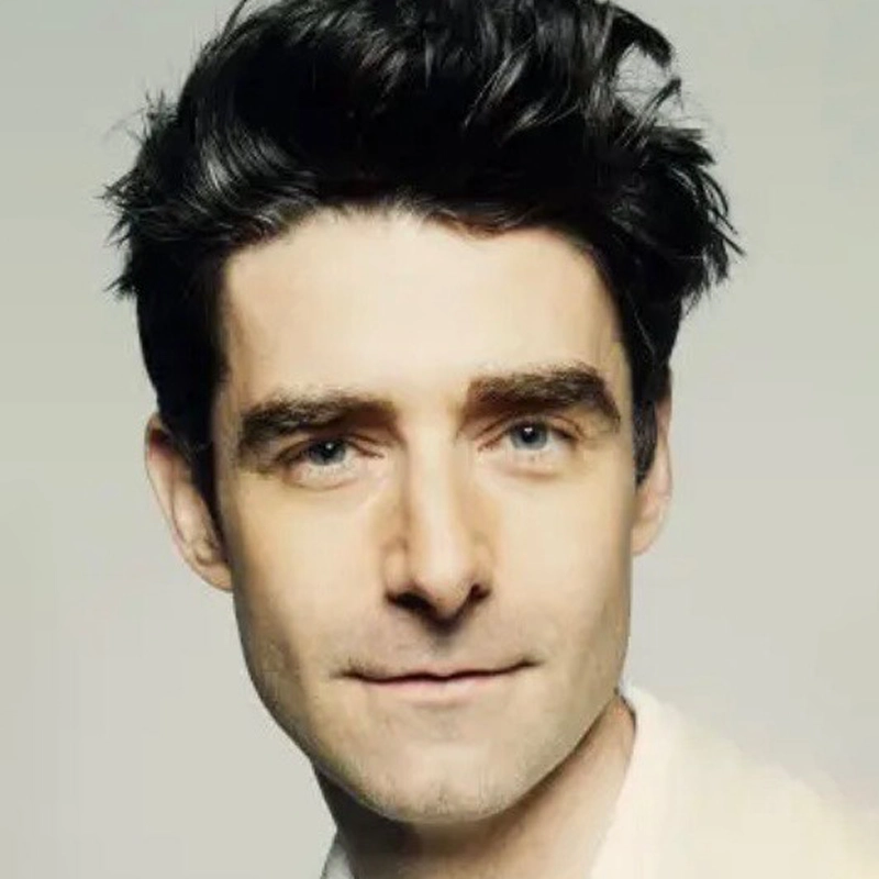 A young man with fair skin and tousled dark hair looks directly at the camera with a subtle, closed-mouth smile. He has blue eyes, strong eyebrows, and a clean-shaven face—bearing a resemblance to Drew Gehling. The background is a plain, light neutral color.