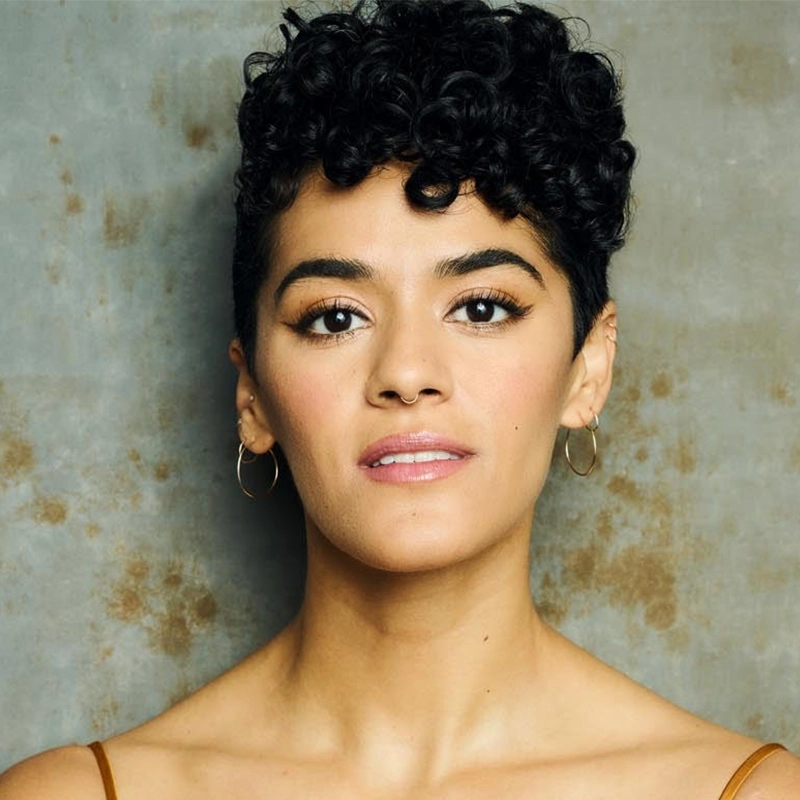 A person with short, curly black hair, gold hoop earrings, and dewy makeup gazes confidently at the camera. They are wearing a thin-strapped top and are posed against a textured, mottled gray-brown background.