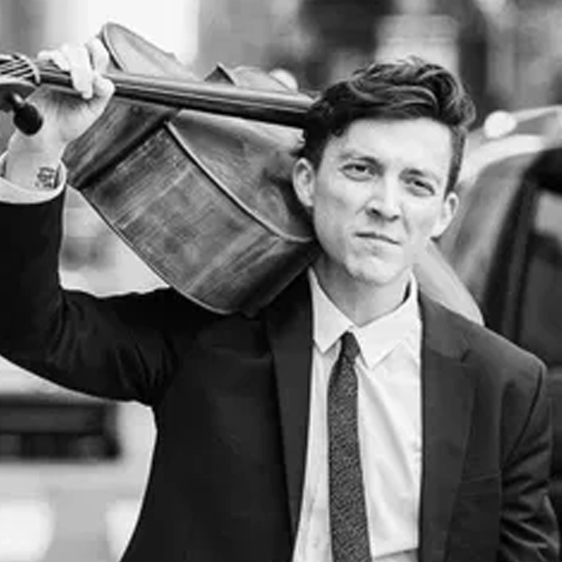 A young man in a suit and tie stands outdoors, holding a cello resting on his shoulder. With short, styled hair and a slight smile, Sam Quiggins is captured in black and white against a blurred city street with vehicles in the background.