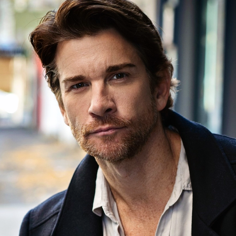 A man with light skin, short brown hair, and a beard looks directly at the camera with a serious expression. He is wearing a dark coat over a light shirt. The background is blurred, showing an outdoor urban setting with muted colors.