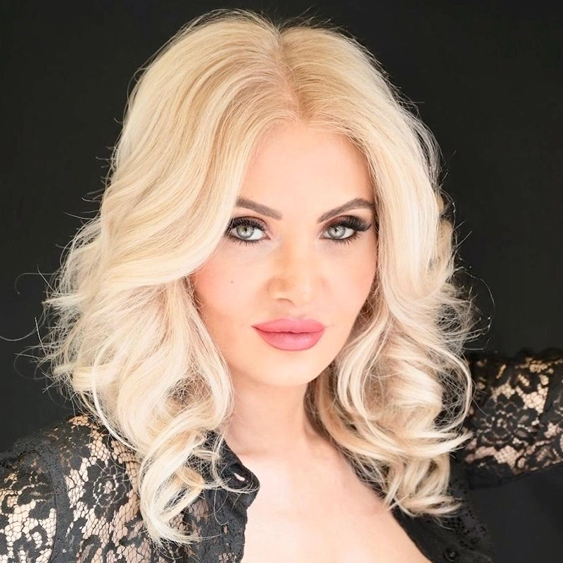 A woman with platinum blonde, wavy hair poses against a black background. She has light skin, striking blue eyes with bold eye makeup, and wears pink lipstick. She is dressed in a black lace top and looks confidently at the camera.