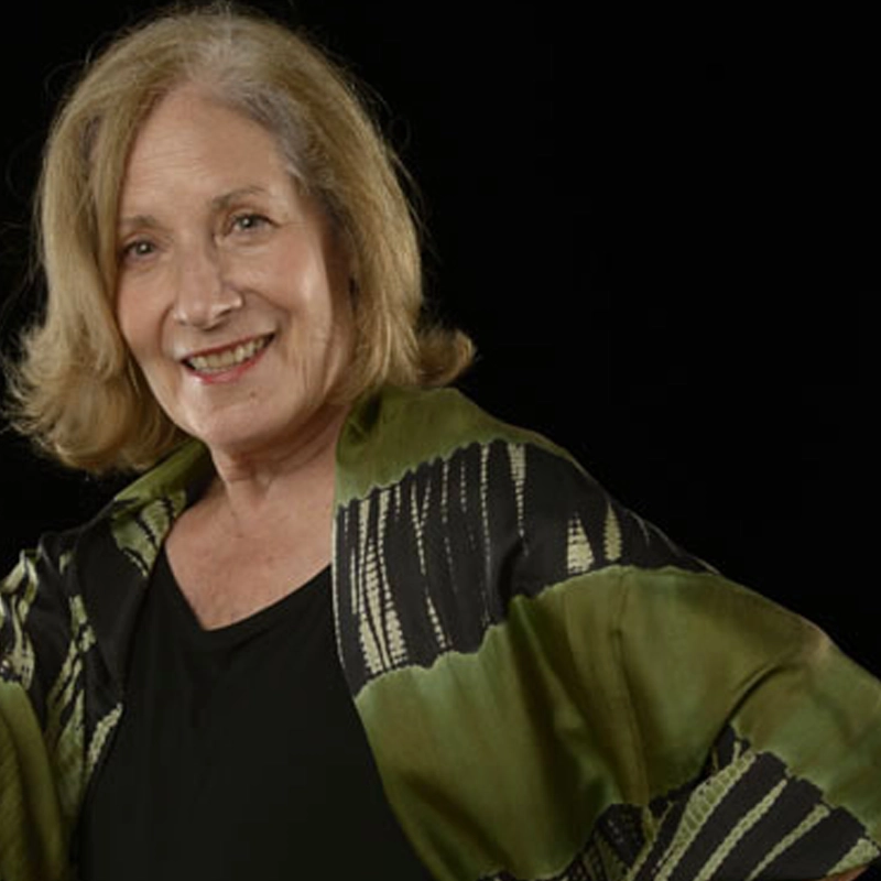 An older woman with shoulder-length blonde hair smiles gently. She wears a black top and a green-black patterned shawl. The background is black, highlighting her warm expression and relaxed pose.