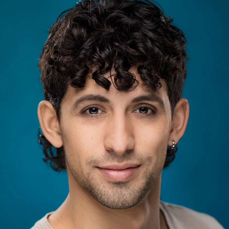 A young man with short, curly dark hair and light facial hair smiles softly. He has light brown skin, dark eyes, and wears a small hoop earring in his left ear. The solid teal blue background highlights Brian Myers Cooper’s relaxed charm.