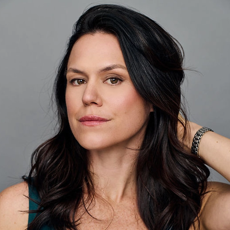 A portrait of Laura D’Andre, a woman with long, dark wavy hair, looks directly at the camera with a neutral expression. She is wearing a dark green top and a silver link bracelet on her left wrist, set against a plain gray background.