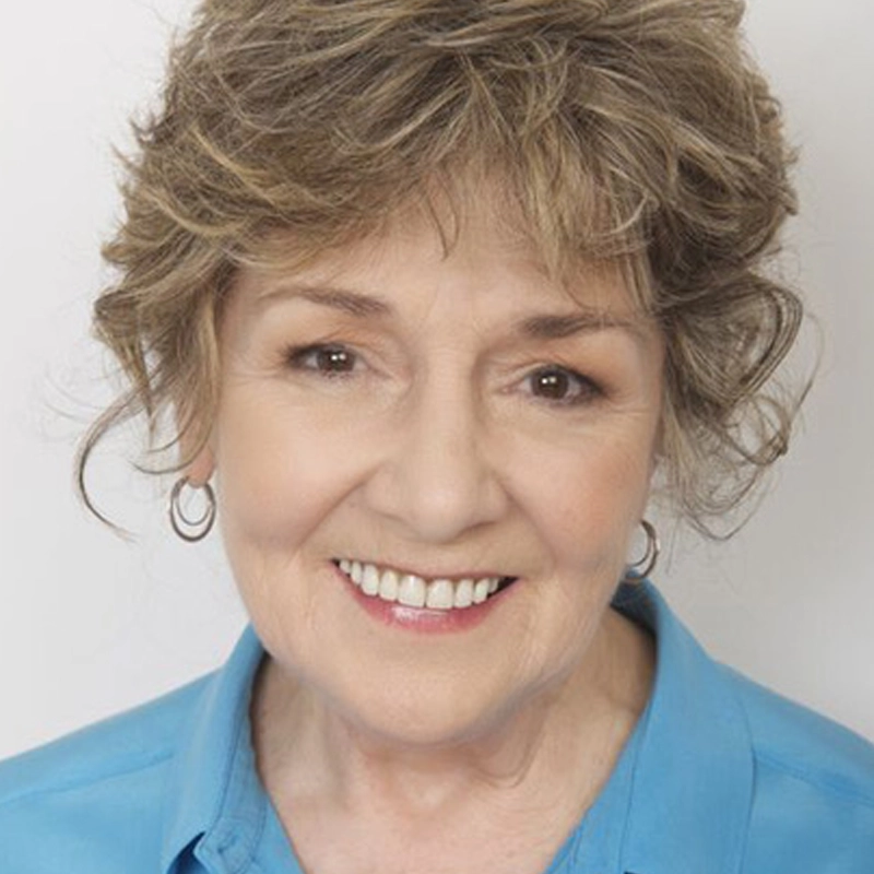 An older woman, Patti Mariano, with short, curly, light brown hair and hoop earrings smiles warmly. She is wearing a light blue collared shirt and is posed in front of a plain, light background. Her expression is friendly and inviting.
