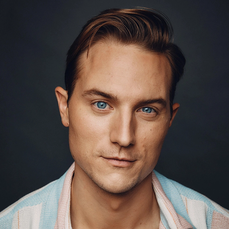 A young man, resembling Brian Myers Cooper, with short, light brown hair styled neatly, blue eyes, and fair skin looks directly at the camera. He wears a light blue and white collared shirt over a light-colored undershirt. The background is dark and plain.