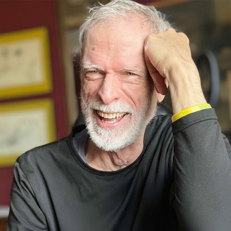 Steve Schalchlin, an older man with white hair and a beard, smiles brightly as he rests his head on his hand. He wears a black shirt and yellow wristband. The softly blurred background reveals framed pictures on a red wall.