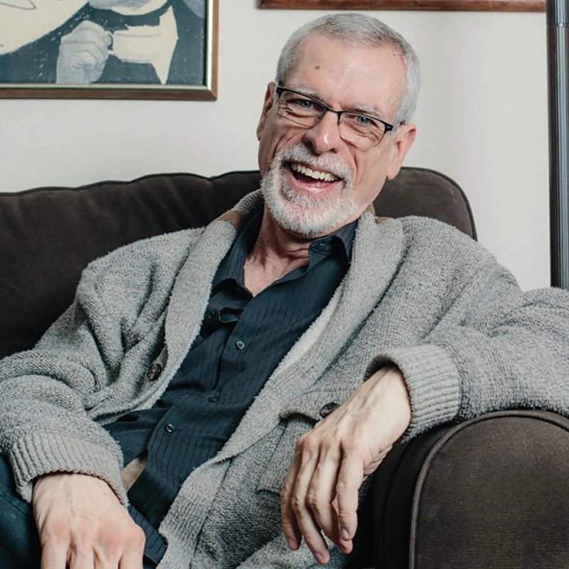 An older man with short gray hair, glasses, and a trimmed beard smiles widely while sitting on a dark couch. Steve Schalchlin wears a gray cardigan over a dark shirt. Framed artwork is visible behind him in the cozy, relaxed setting.