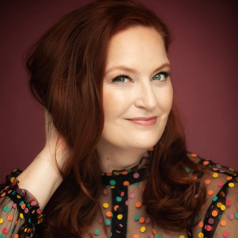 Lexie Dorsett Sharp, with long red hair, smiles softly at the camera. She wears a sheer black top adorned with colorful polka dots, gently touching her hair. A warm burgundy background and soft lighting highlight her features beautifully.