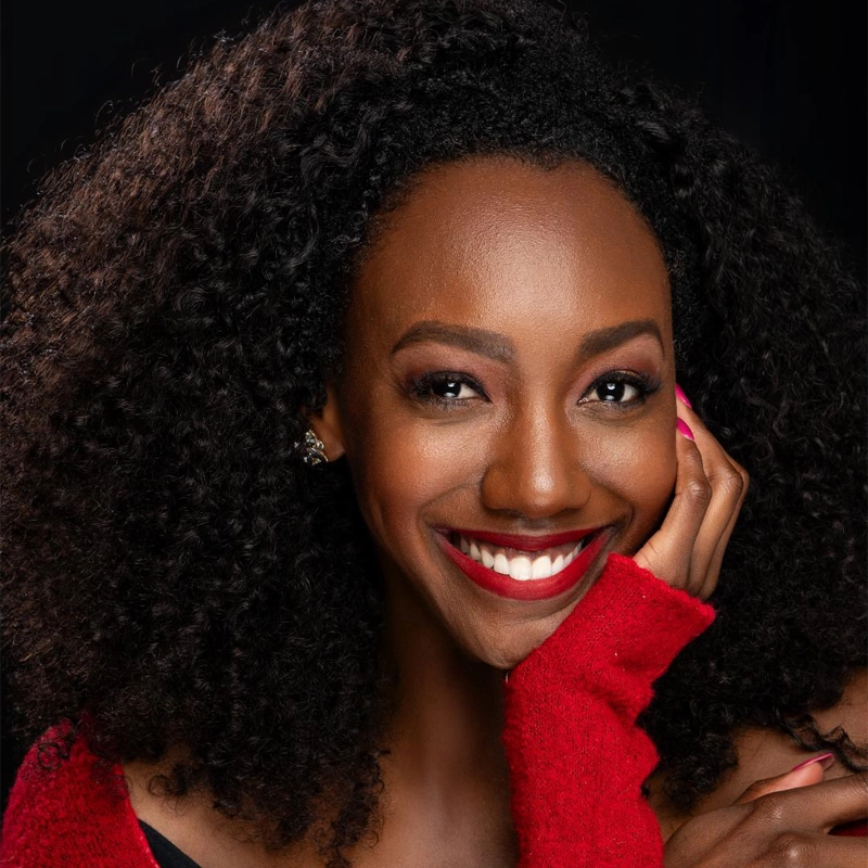 Zurin Villanueva, a woman with dark curly hair, smiles warmly. She wears a red sweater, pink nail polish, small earrings, and red lipstick. Her hand rests gently on her cheek against a black background. Her expression is joyful and confident.