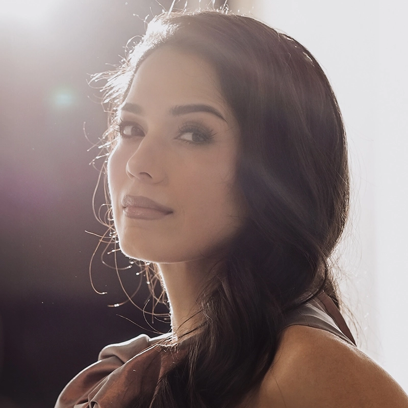Actress Arielle Jacobs looks toward the camera with long, dark hair, her face softly illuminated by a bright backlight. She has a neutral expression and wears a muted, ruffled garment, with a lens flare appearing in the upper left corner.