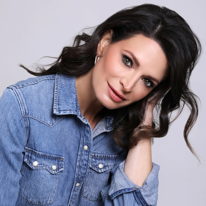 Jackie Burns, with wavy dark hair and light eyes, tilts her head, smiling softly. She wears a blue denim button-up shirt with white snaps and silver hoop earrings. Her hand touches her hair against a plain soft gray background.