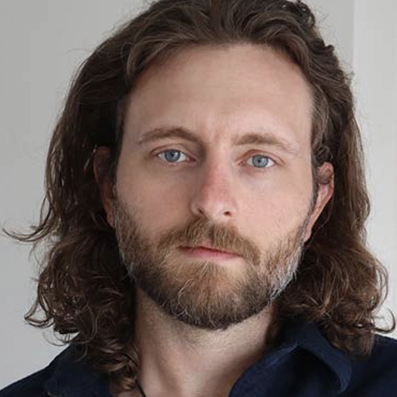 A man with long wavy brown hair, a full beard, and blue eyes looks directly at the camera. He has fair skin and is wearing a dark shirt. The background is plain and light-colored, drawing attention to his thoughtful expression.