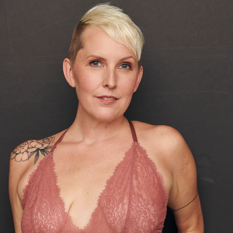 A person with short, platinum blonde hair and light skin poses against a plain dark background. Wearing a mauve lace halter top, they reveal a floral tattoo on their left shoulder—capturing Natalie Joy Johnson's calm, confident gaze directly at the camera.