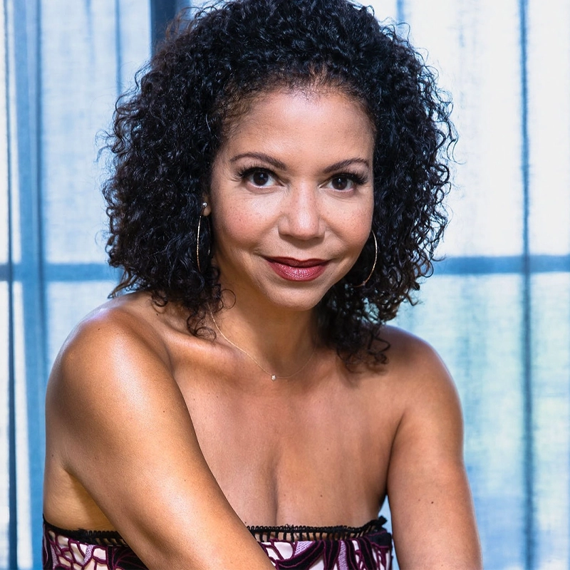 A close-up portrait of singer and actress Gloria Reuben with dark curly hair and a warm smile. She wears a strapless dress with a patterned neckline and a delicate necklace.