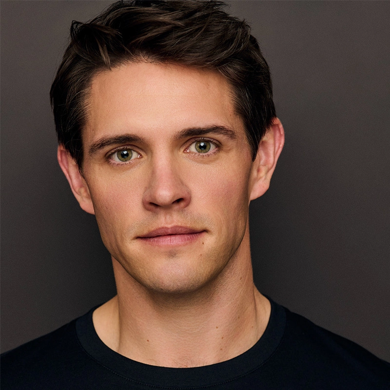 A young man with short dark brown hair and hazel eyes faces the camera against a plain dark background. He has a slight smile and wears a black crew-neck shirt. The lighting is even, highlighting his facial features clearly.