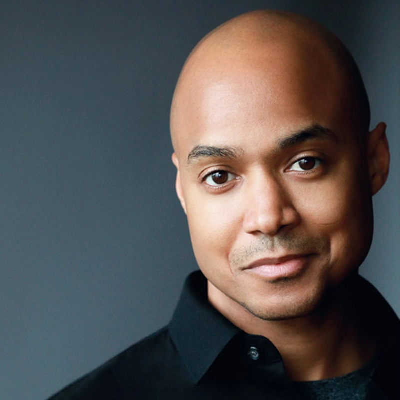 Curtis Wiley, a bald man with medium brown skin and dark eyes, smiles softly. He is wearing a black collared shirt, with a smooth grey gradient background that gives the photo a professional and friendly appearance.