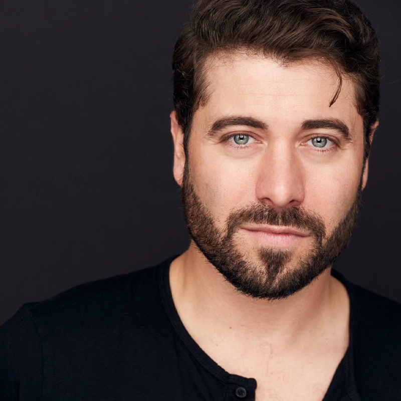 Omar Lopez-Cepero, with light skin, short dark hair, and a trimmed beard, gazes confidently at the camera. He wears a black shirt with a few buttons undone; the plain dark background highlights his bright eyes and gentle expression.