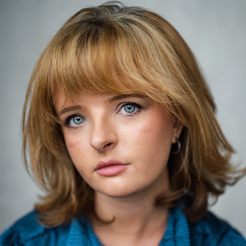 A headshot of actress Milly Shapiro with shoulder-length, honey-blonde hair and light blue eyes looking directly at the camera. She has a neutral expression and is wearing a dark teal, textured shirt against a plain, light grey background.