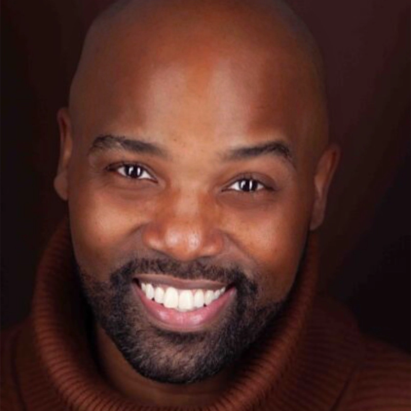 A smiling Melvin Tunstall III, bald with dark skin, neatly groomed beard and mustache, wears a brown turtleneck sweater. He faces the camera against a soft, dark background, showing white teeth and joyful, expressive eyes.