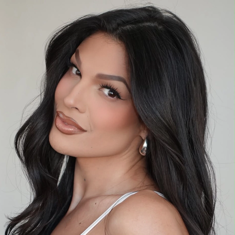 Portrait of actress Milly Shapiro with long, wavy black hair and glamorous makeup, including winged eyeliner and glossy brown lips. She is wearing small silver hoop earrings and a white top against a neutral background, looking directly at the camera with a slight smile.