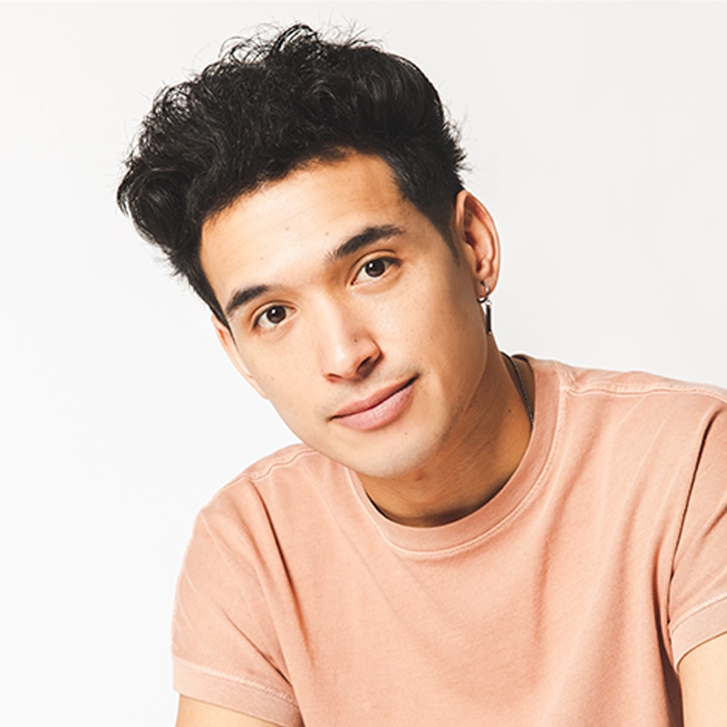 A young person with short, curly dark hair and light skin is smiling gently, facing forward. Wearing a light pink t-shirt and a small hoop earring, they evoke Zachary Noah Piser’s signature style against a plain white, bright background.