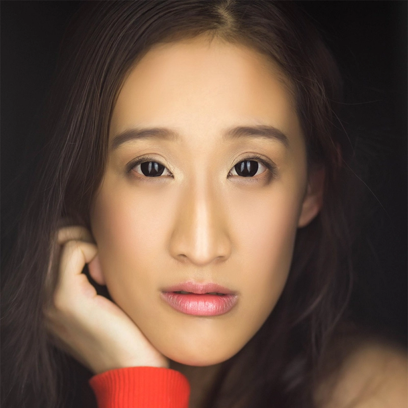 A young woman with long, dark hair gazes directly at the camera. Her face is softly lit, highlighting smooth skin and natural makeup. She rests her chin on her hand, wearing a bright orange sleeve—captured in the signature style of Mitchell Nalos.