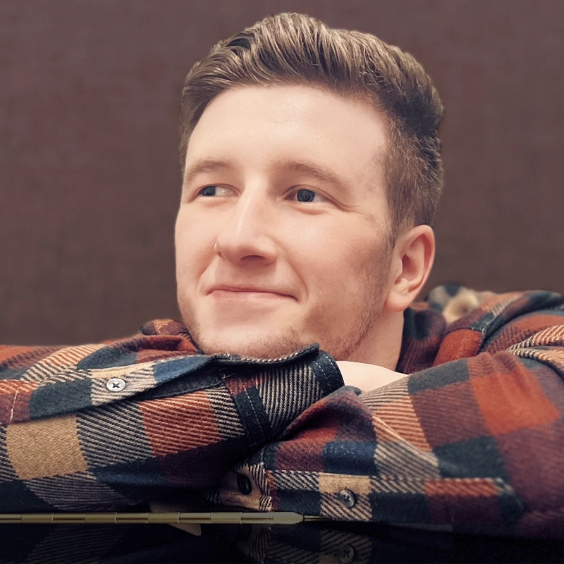Zach Anderson, a young man with short, light brown hair and a trimmed beard, rests his arms on a surface, smiling softly. He wears a red, black, and beige plaid flannel shirt against a plain brown background for a warm, relaxed feel.