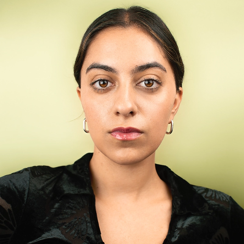 A woman, resembling Nadia Vito, with dark hair parted in the middle, wearing a black top and silver hoop earrings, looks directly at the camera with a neutral expression. The background is a solid light green color.