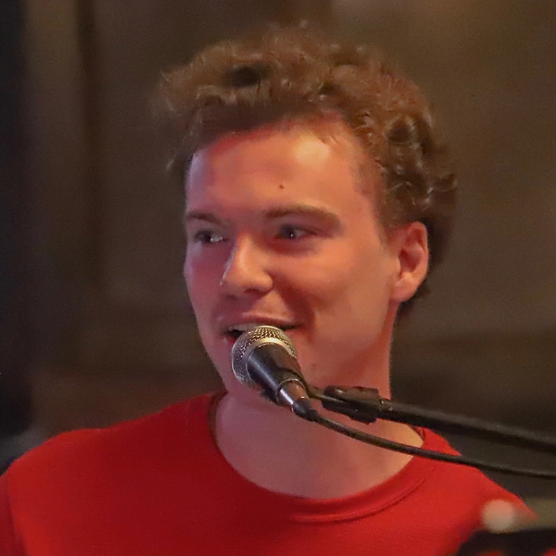 A young man with curly hair wearing a red shirt speaks or sings into a microphone. The softly blurred background draws focus to Vito’s smiling expression and the dim, warm indoor lighting.