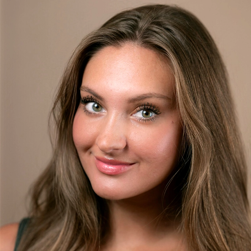 A young woman, Nadia Jewel, with long, light brown hair and light eyes smiles softly. She has smooth, glowing skin, natural makeup, and wears a dark top. The beige background is plain and the image is closely cropped to her face and shoulders.