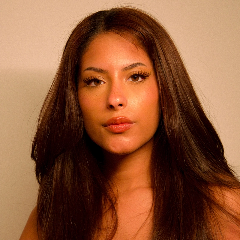 A young woman, Nadia Jewel, with long, straight brown hair and warm-toned skin looks directly at the camera with a neutral expression. She has full lips, defined eyebrows, and wears natural makeup against a softly lit plain background.