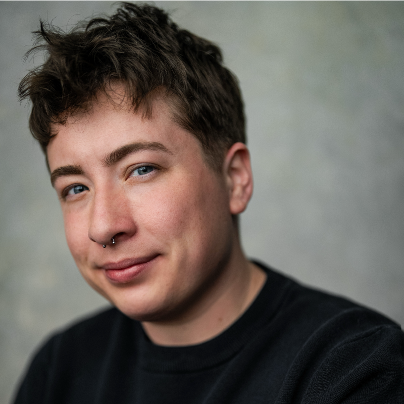 Kathel Griffin, with short, tousled brown hair, blue eyes, and a nose ring, smiles gently. They are wearing a black sweatshirt and are posed against a softly blurred, neutral background.