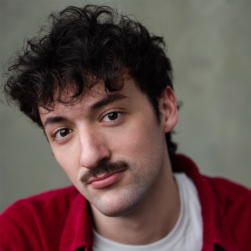A young man with curly dark hair and a mustache looks into the camera with a slight smile. He wears a red shirt over a white t-shirt, reminiscent of Vito, and the background is softly blurred in neutral tones.