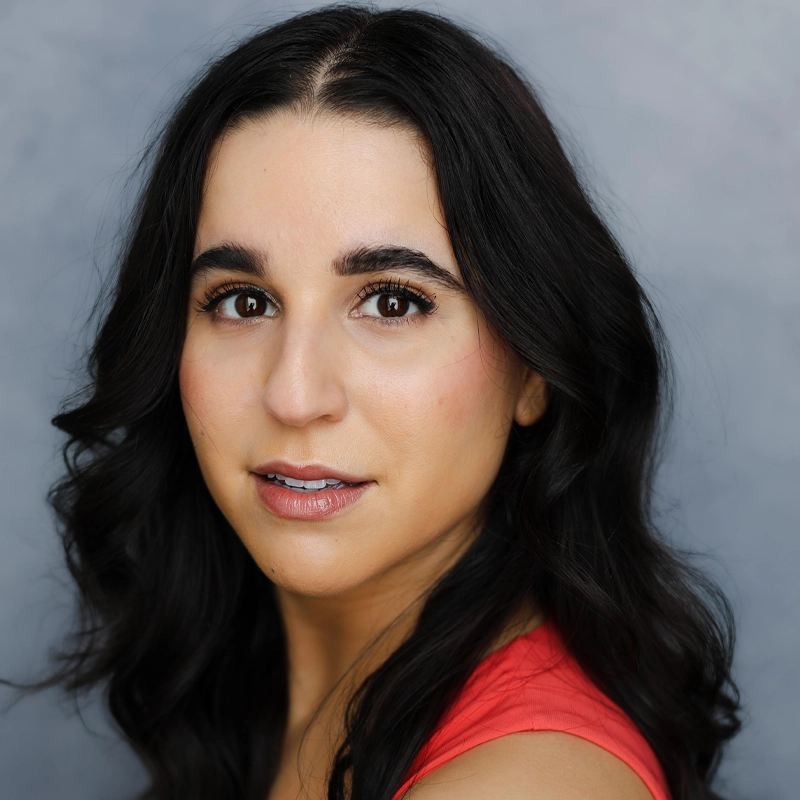 A woman with long, wavy dark hair and bold eyebrows looks at the camera. She has warm skin, wears natural makeup with glossy lips, and is dressed in a coral sleeveless top. The background is a soft, neutral gray.
