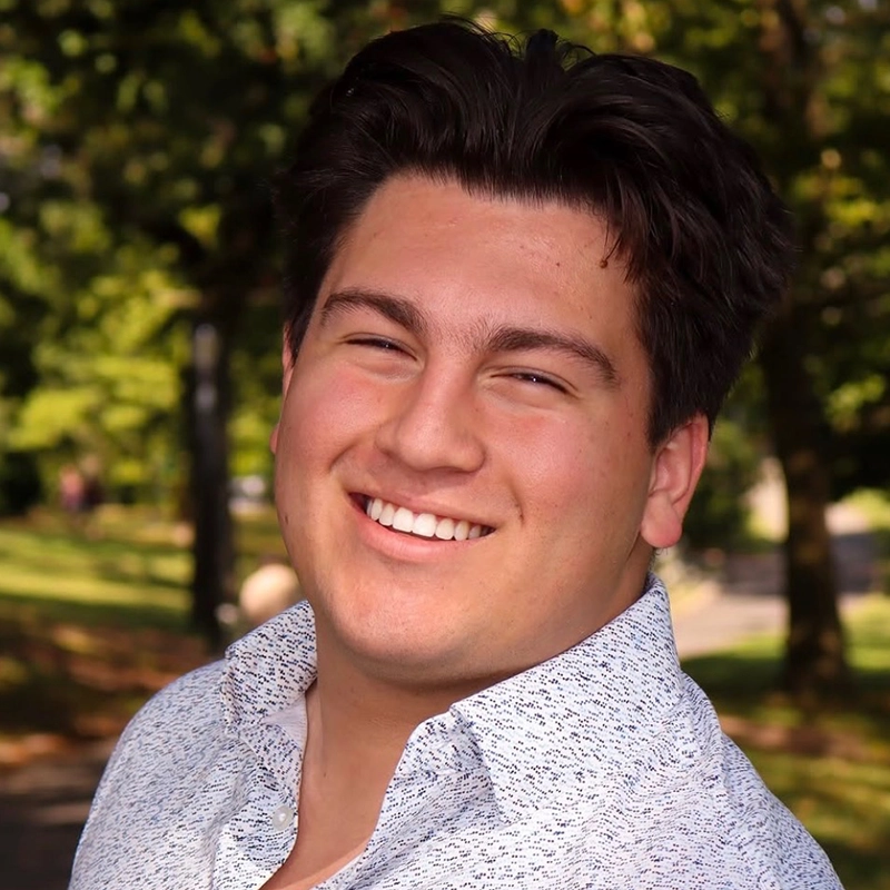 A young man with dark, wavy hair smiles warmly at the camera. Vito is wearing a light-colored, patterned button-down shirt and standing outdoors, with trees and greenery softly blurred in the background. Sunlight highlights his face.