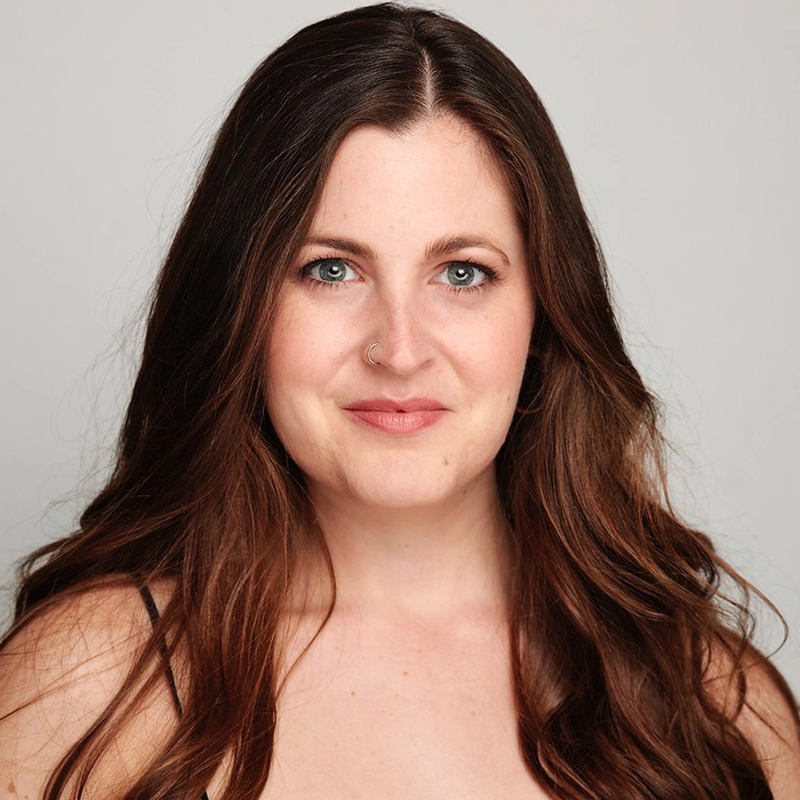 A woman with long, wavy brown hair and blue eyes smiles softly at the camera. She has fair skin, wears natural makeup, and has a nose ring. The background is plain and light gray.