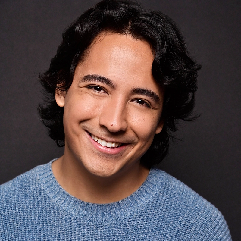 A young person with medium-length wavy dark hair smiles warmly at the camera. Mitchell Nalos wears a light blue textured sweater and is posed against a plain dark background, creating a friendly and approachable portrait.
