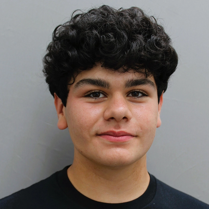 A young person, possibly named Vito, with short, curly dark hair and thick eyebrows looks at the camera with a neutral expression. They have medium skin, wear a black crew-neck shirt, and stand against a plain light gray background.