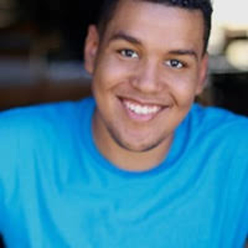 Norberto Troncoso, a young man with short, curly dark hair and medium skin tone, smiles at the camera in a bright blue shirt. The background is blurred, drawing focus to his friendly expression.