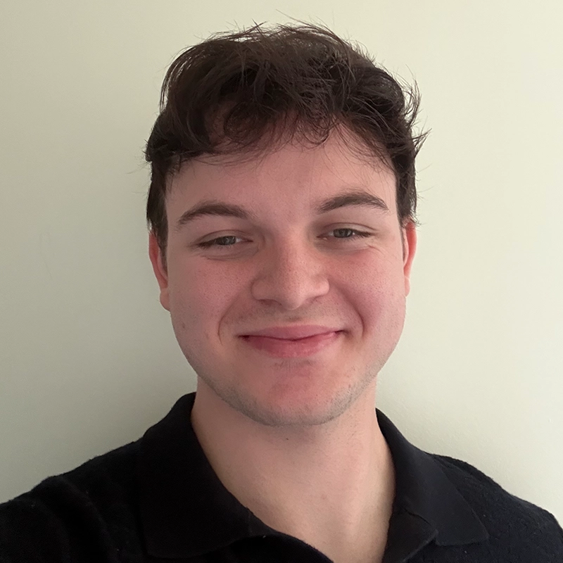 A young person with fair skin, short dark brown hair, and hazel eyes smiles softly at the camera. Wearing a black collared shirt, Vito is posed against a plain, light-colored background.