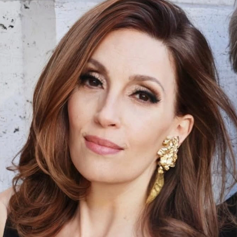 Woman with wavy brown hair, gold floral earrings, and natural makeup smiles slightly. She is wearing a black top and is photographed against a light-colored textured background. Only her face and upper shoulder are visible, with soft lighting highlighting her features, reminiscent of Barbara Fasano’s signature elegance.
