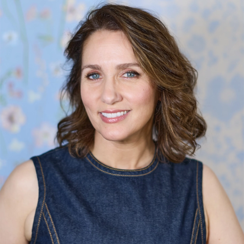 Tricia Paoluccio, a woman with wavy brown hair and blue eyes, smiles gently. She wears a dark denim sleeveless top with tan stitching. The background is pale blue with a subtle, floral pattern. The lighting is soft, highlighting her natural features.