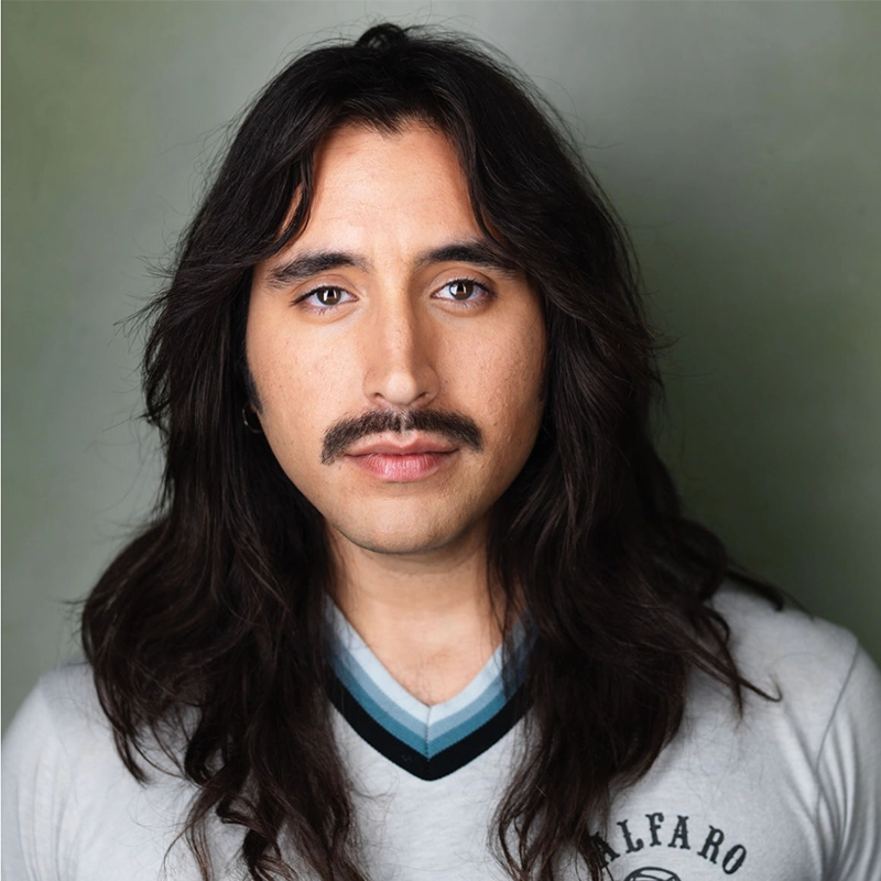 Anthony Alfaro, with long, wavy dark hair and a mustache, looks directly at the camera. He wears a light gray V-neck shirt with dark blue and white stripes on the collar. The background is a plain, muted green.