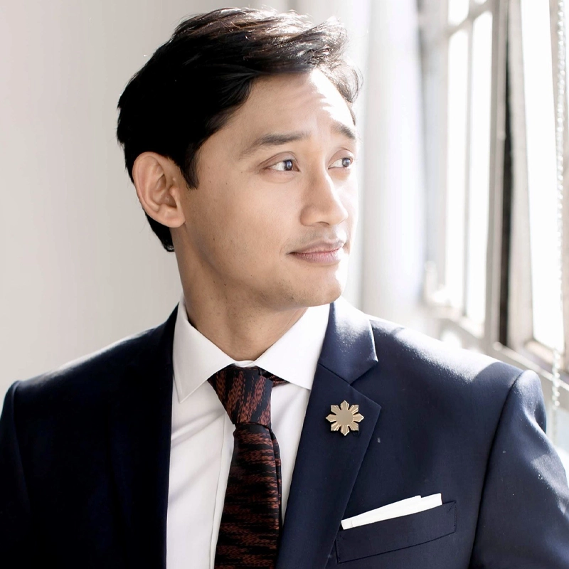 A well-groomed man, resembling Joshua Dela Cruz, in a navy suit, white shirt, and patterned tie stands indoors near a sunlit window. He looks pensively to the side, with a gold flower-shaped pin and a white pocket square on his suit.