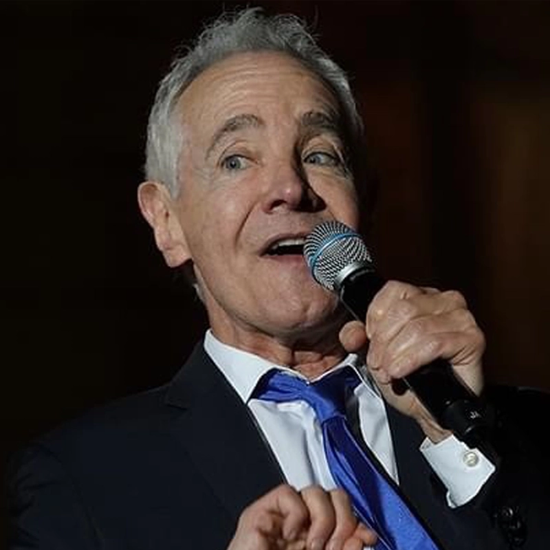 An older man with gray hair, wearing a dark suit, white shirt, and blue tie, holds a microphone close to his mouth while speaking or singing. He is looking slightly upward, with an expressive face, against a dark, blurred background.